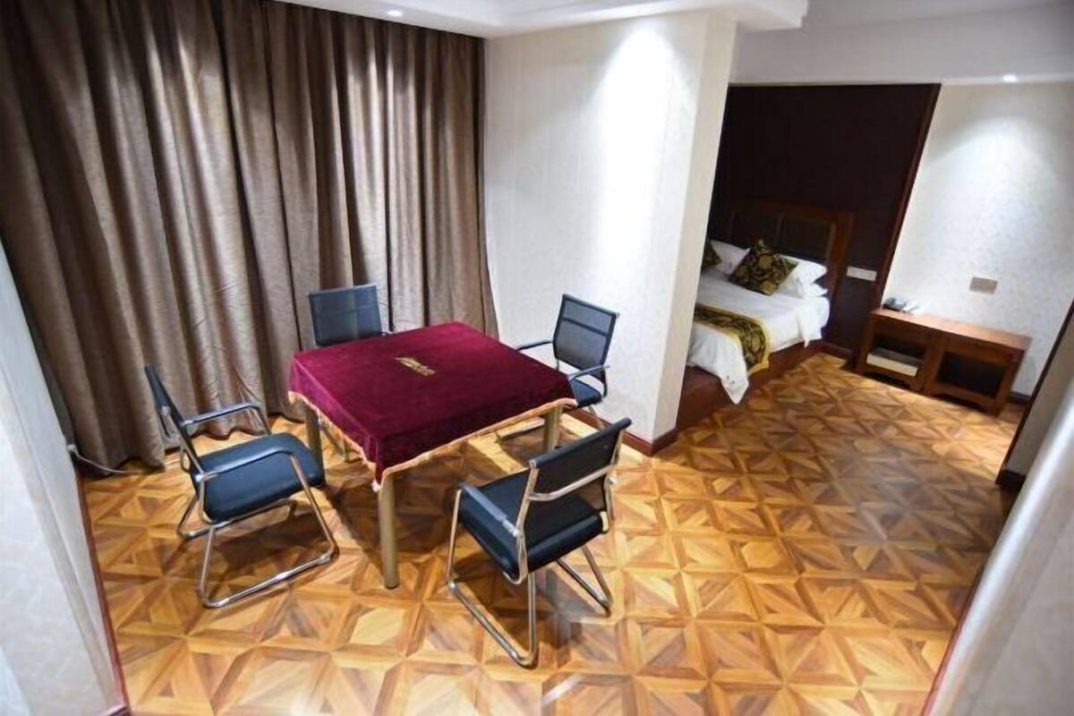 A hotel room with a wooden parquet floor, a red table with four chairs, a bed with a white comforter, and a nightstand with a lamp.
