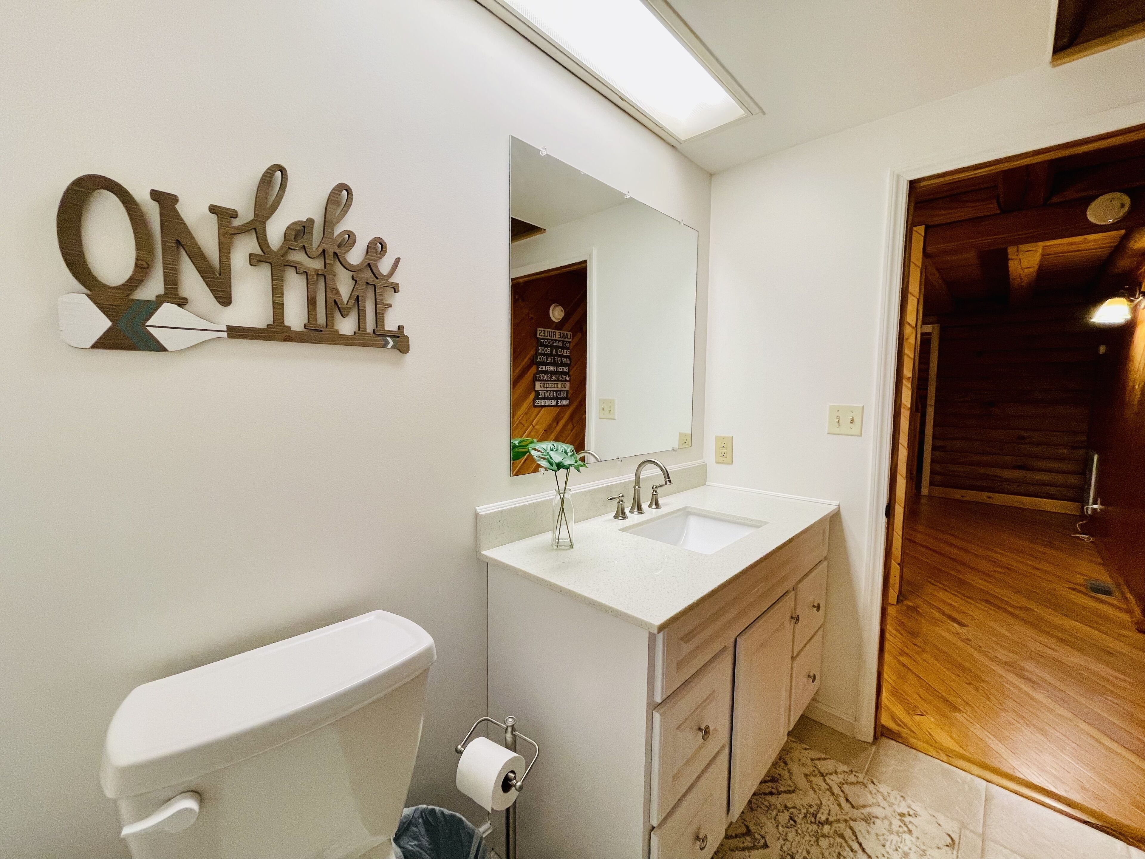 Un baño con inodoro blanco, un lavamanos con espejo y una puerta de madera que da a otra habitación.