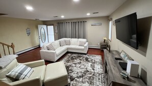 Ein Wohnzimmer mit Ecksofa, einem kleinen Tisch, einem Flachbildfernseher und einem großen Fenster mit Vorhängen.