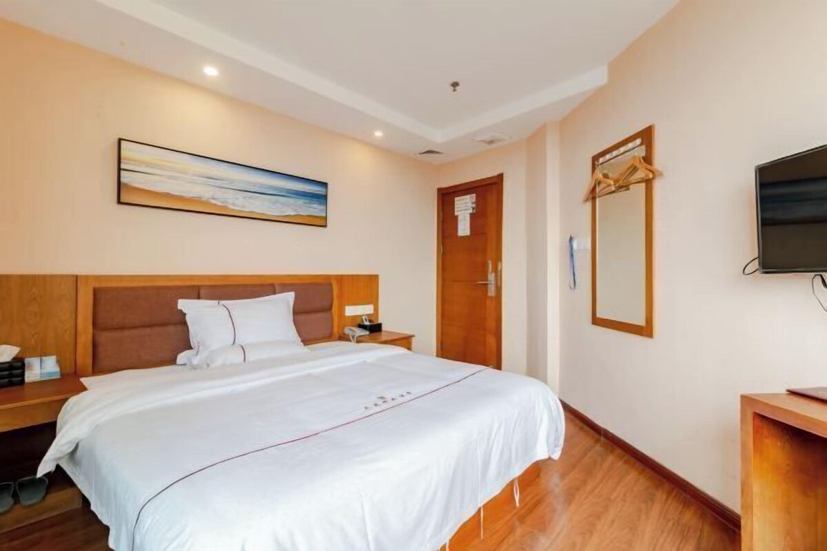 Habitación de hotel con una cama grande, cabecero de madera, un cuadro en la pared, una puerta, un espejo y un televisor sobre un mueble de madera.