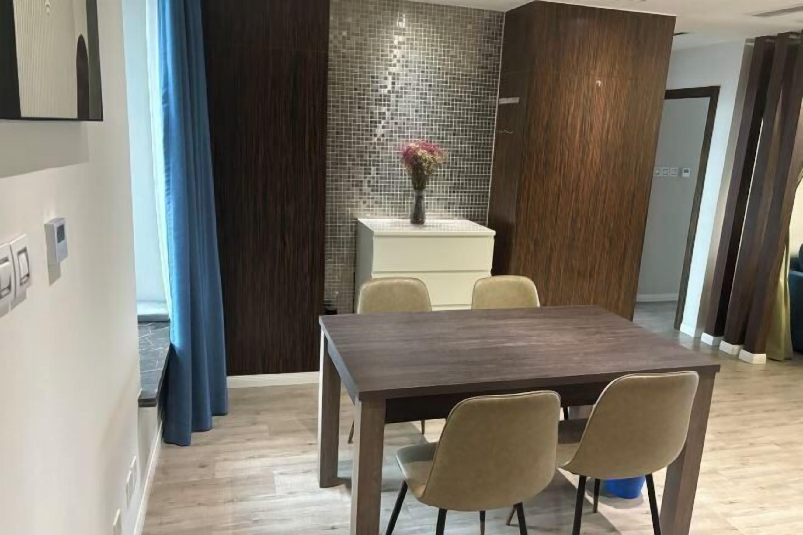 Un comedor moderno con una mesa de madera oscura y sillas color beige, un mueble blanco con un jarrón de flores y un salpicadero de azulejos.