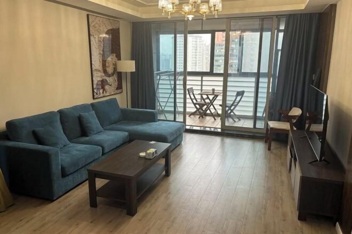 A modern living room with a sofa, coffee table, and a TV stand. There is a balcony with a table and chairs.