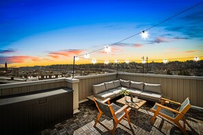 Terraza en la azotea con sofá, sillas y una mesa, con vistas a un paisaje urbano al atardecer.
