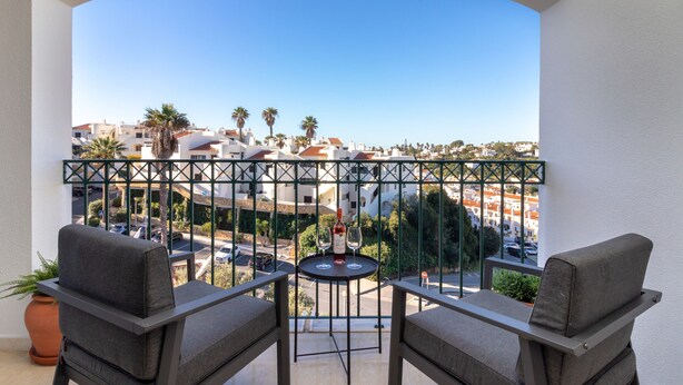 A balcony with two grey sofas, a small round table with two wine glasses and a bottle, and a view of a cityscape with buildings and palm trees. - Casa Encantada - Communal pool & garden, just a 5 minute walk to town centre (Carvoeiro)