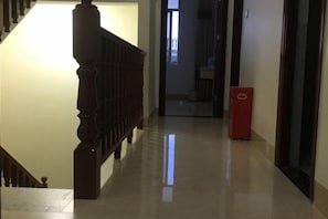 A hallway with a wooden staircase on the left, a red trash bin on the right, and a door at the end.