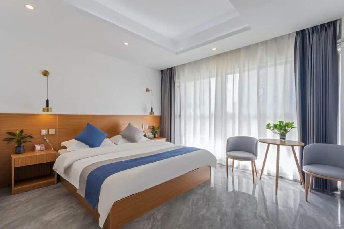 Een moderne hotelkamer met een houten hoofdeinde, een groot bed met blauwe accenten, een nachtkastje met een plant, een ronde tafel met twee stoelen en een groot raam met gordijnen.