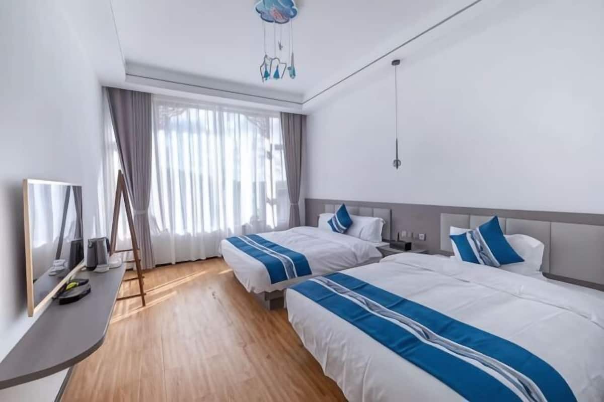 A hotel room with two beds, a desk, a TV, and a large window with curtains.