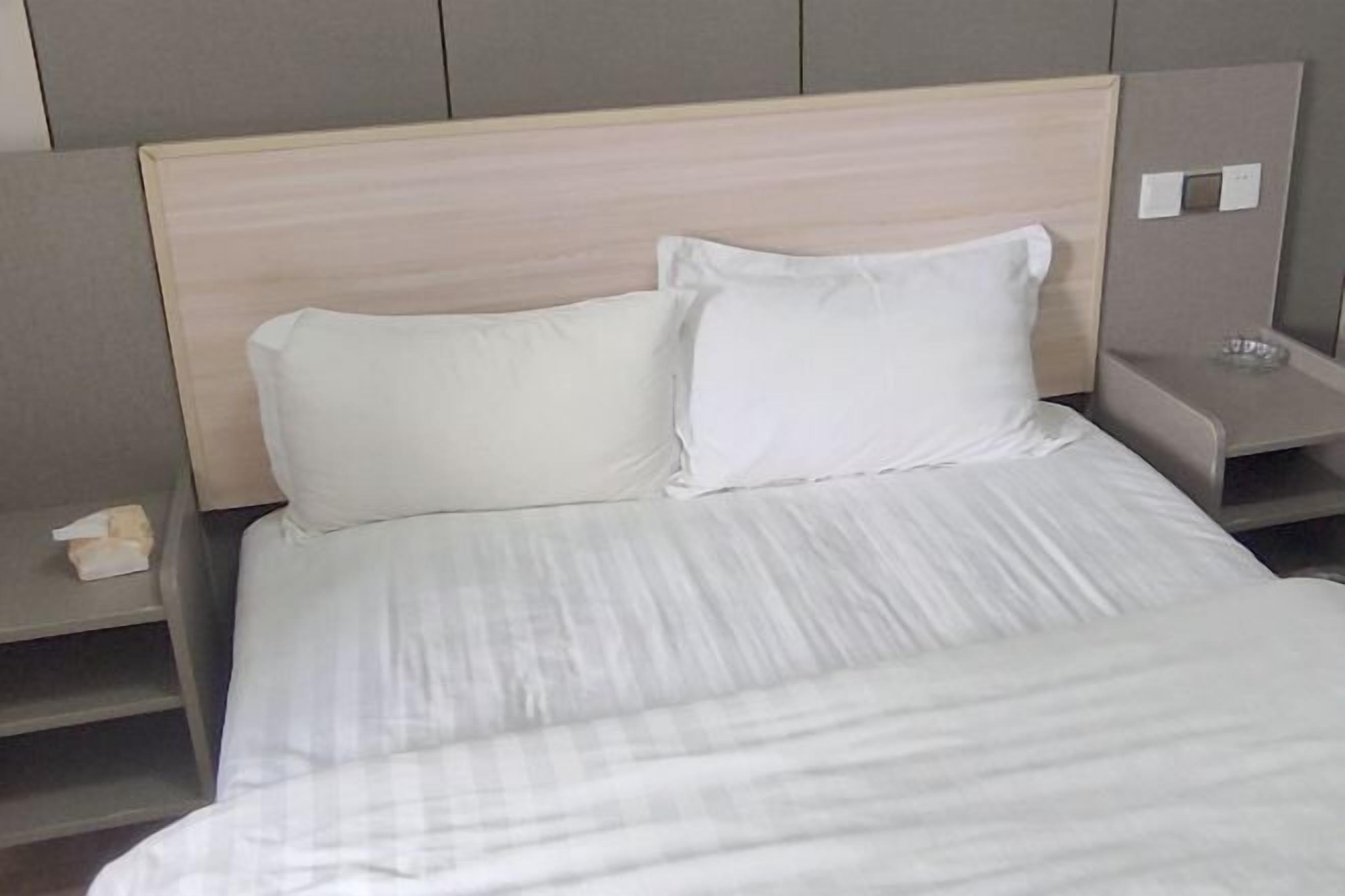 Un lit bien fait, avec du linge de lit blanc et des oreillers, une table de chevet sur laquelle se trouve une boîte de mouchoirs, et un interrupteur fixé au mur.