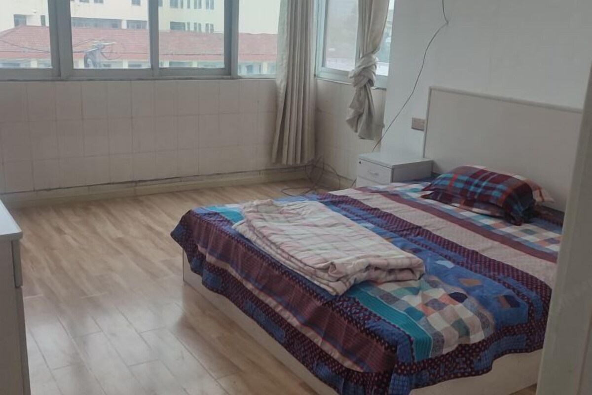A bedroom with a bed, a nightstand, a window with curtains, and a view of buildings outside.