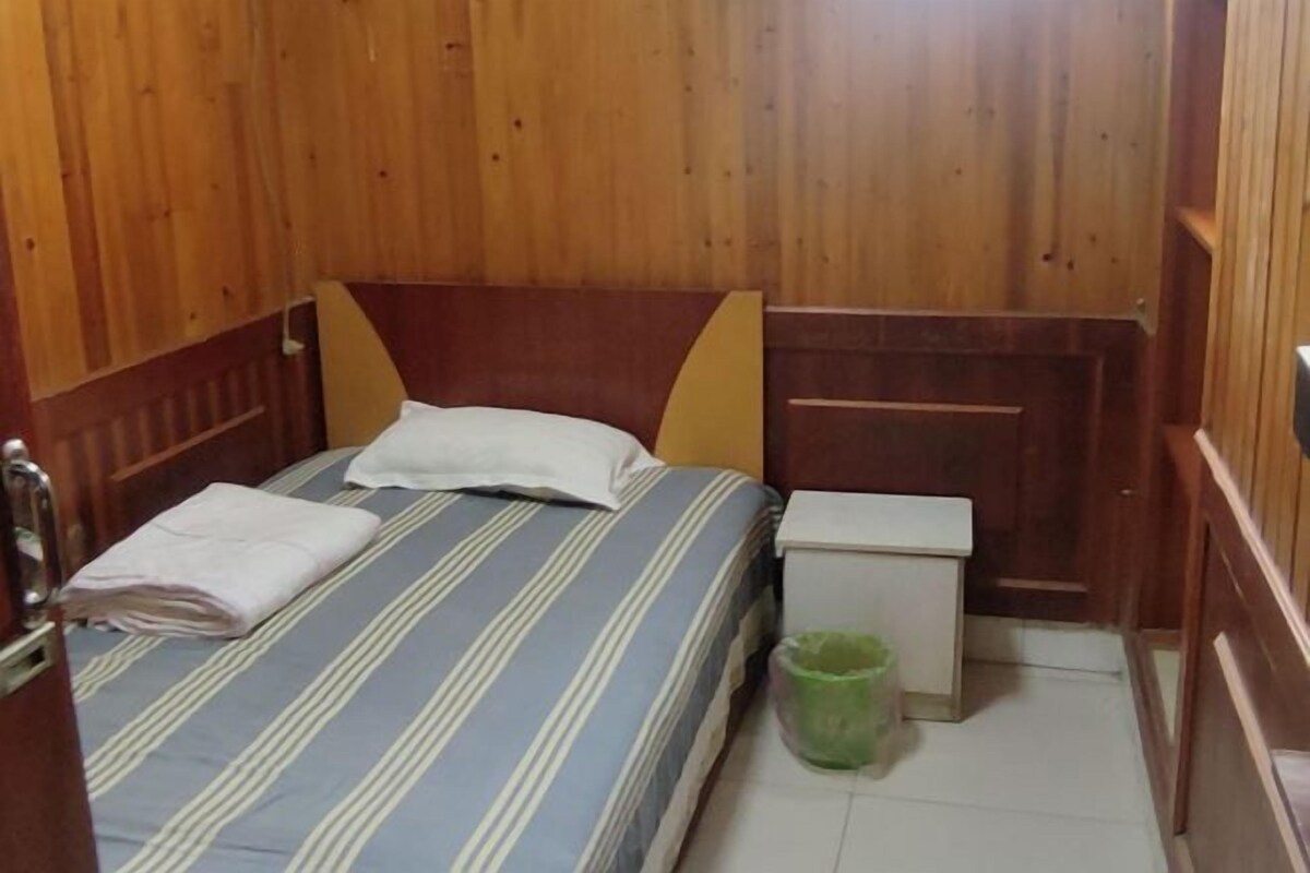 A small, compact room with a single bed, a bedside table, and a green trash bin.