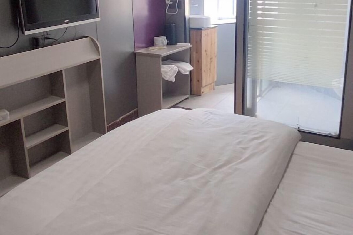 A hotel room with a bed, a TV on a stand, a wardrobe, and a window with blinds.