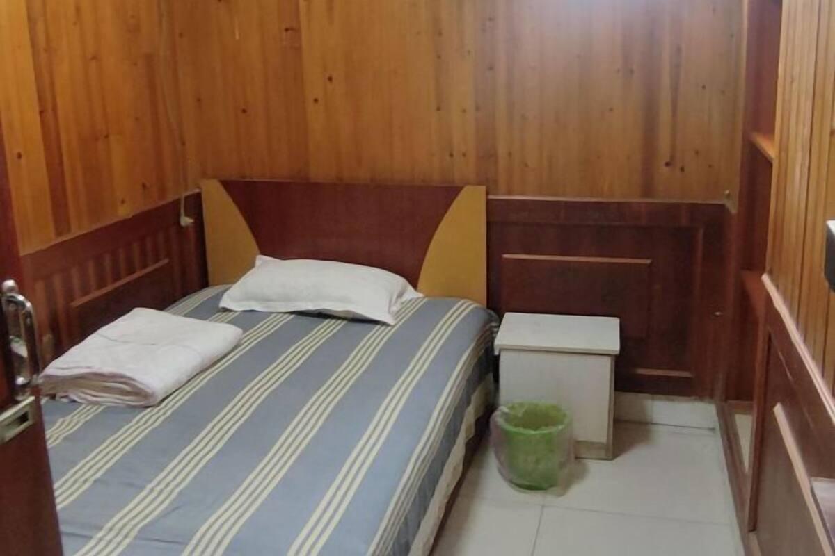 A small, wooden-paneled room with a single bed, a nightstand, and a green trash bin.