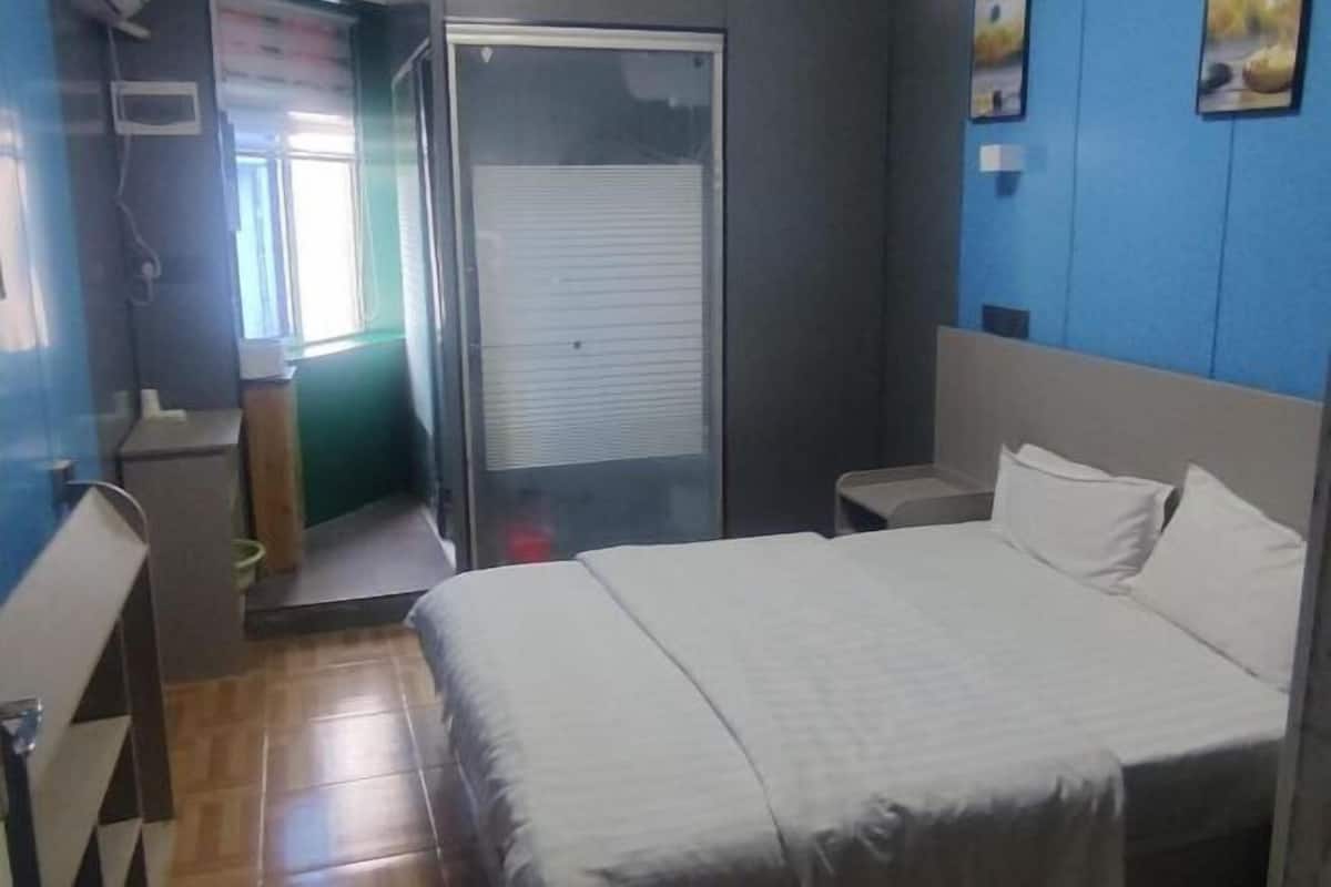 A hotel room with a bed, a small bedside table, a window with blinds, and a door leading to a hallway.