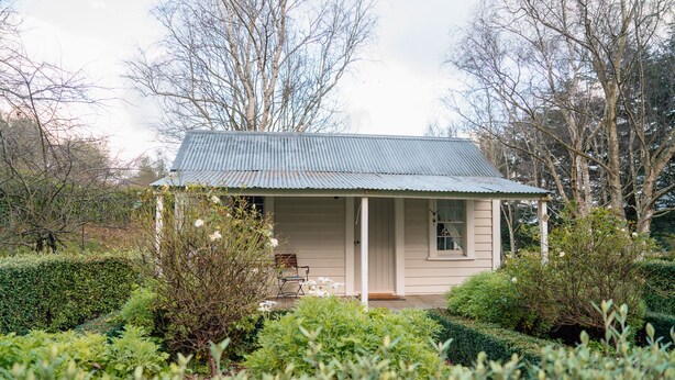 A small, single-story house with a metal roof, surrounded by well-maintained hedges and trees. - Tuhitarata Hideaway. Romantic Couples Retreat (Pirinoa)