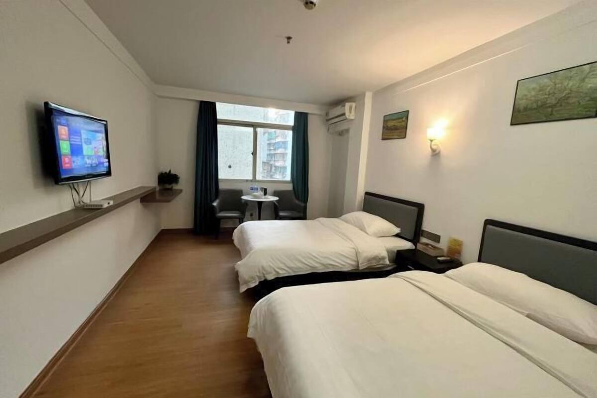 A hotel room with two beds, a TV on the wall, a window with curtains, and a small table with chairs.