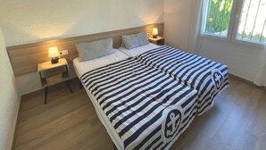 A double bed with striped bedding, two bedside tables with lamps, a window with curtains, and a wooden floor. - Las flores 3A, Begur, Costa Brava (Begur)