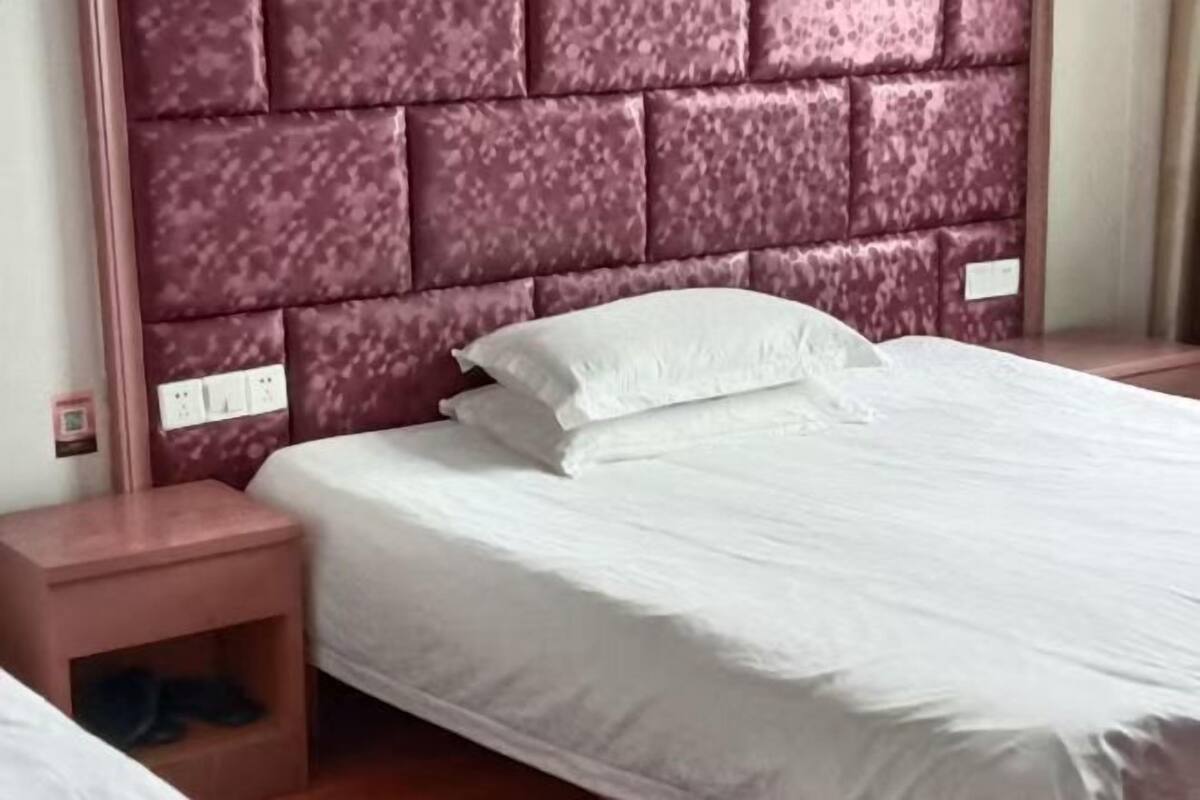 A hotel room with a padded headboard, a single bed with white bedding, a nightstand with a shoe rack, and a power outlet.