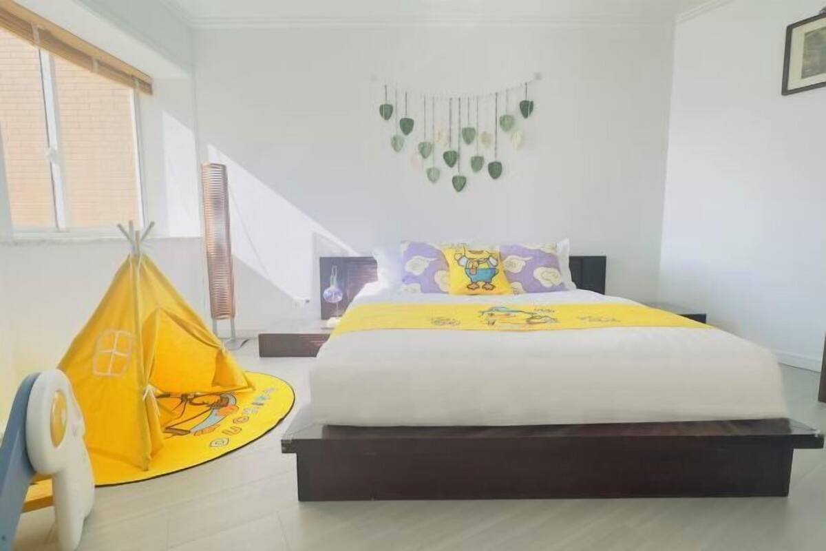 Une chambre avec un lit, une tente de jeu jaune, une décoration murale et une fenêtre avec des stores.