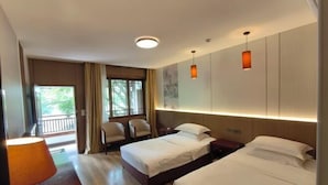 Quarto de hotel com duas camas, área de estar com cadeiras, uma janela grande com cortinas e uma luminária.