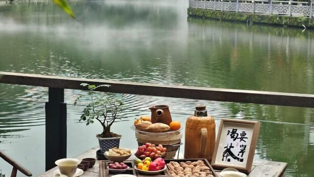 Uma mesa com diversos alimentos e um bule, com um lago calmo e uma cerca tradicional ao fundo.