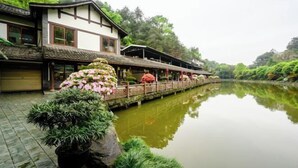 Um edifício tradicional em estilo japonês com um deck de madeira que se estende sobre um rio calmo, rodeado por vegetação exuberante.