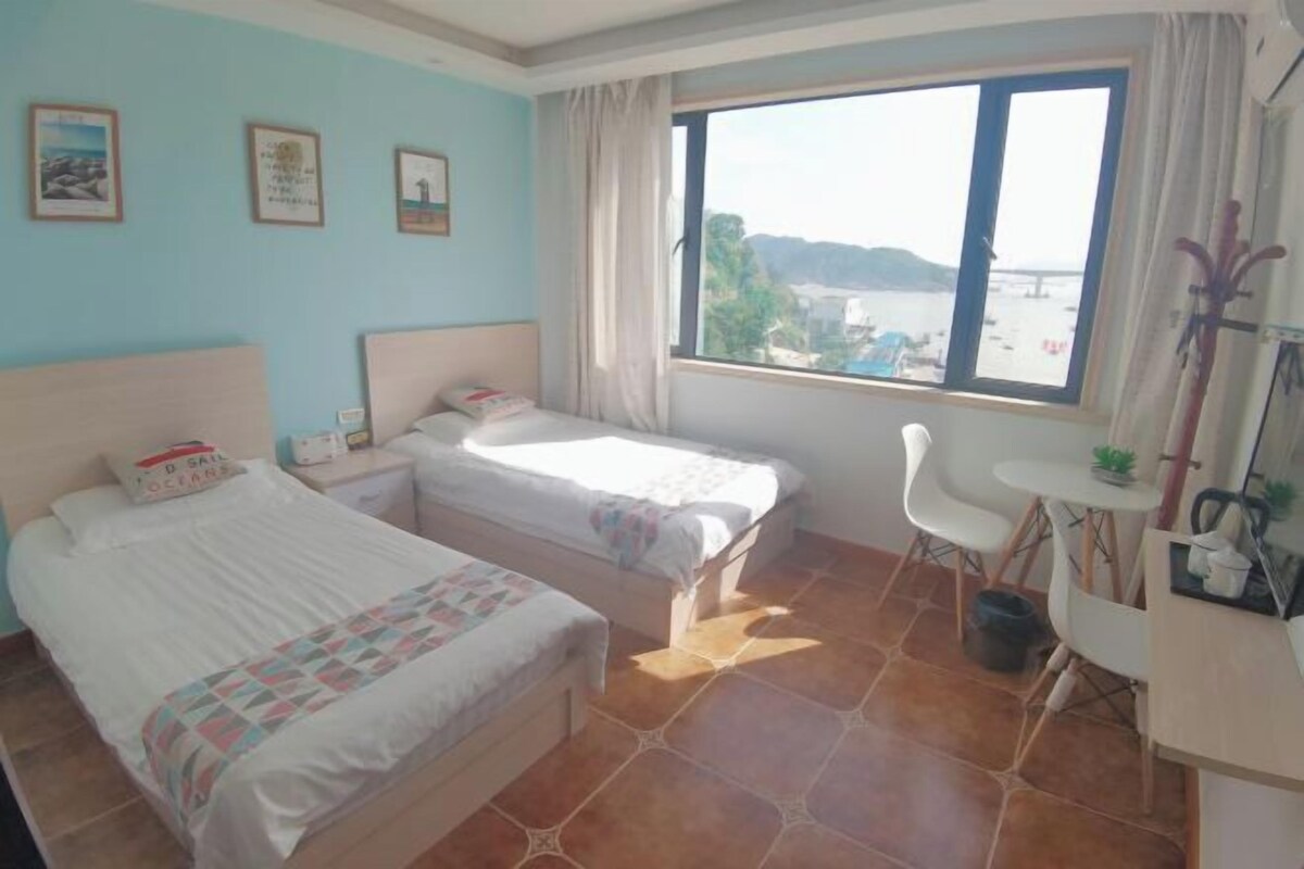 Una habitación con dos camas, una mesita, una silla y una ventana que ofrece vistas a la playa y las montañas.