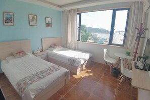 Una habitación con dos camas, una mesita, una silla y una ventana que ofrece vistas a la playa y las montañas.