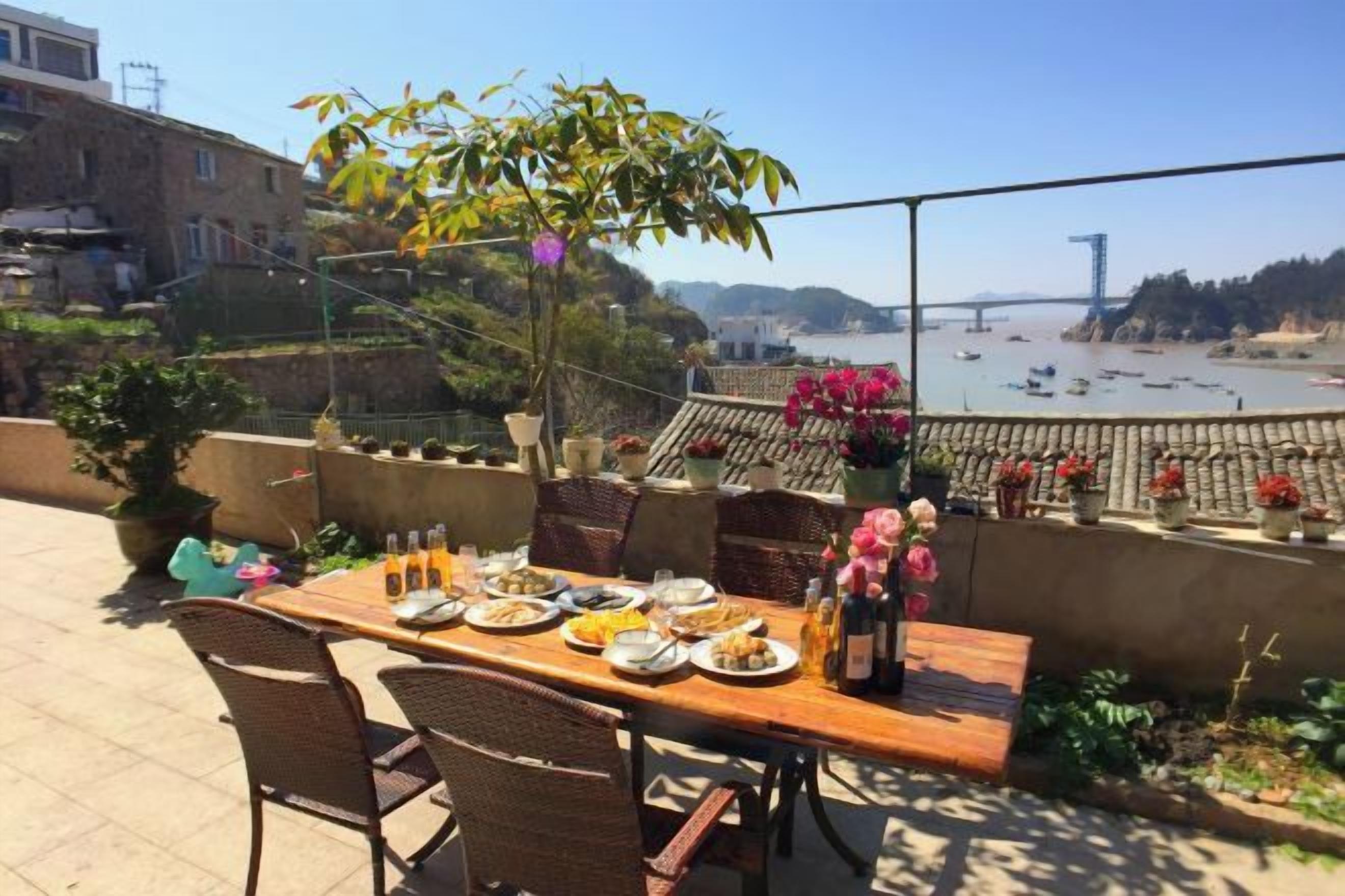 An outdoor dining area with a table set for two, overlooking a beach with boats and a distant view of a bridge.