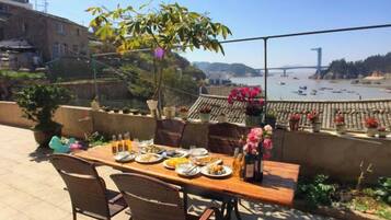 An outdoor dining area with a table set for two, overlooking a beach with boats and a distant view of a bridge.
