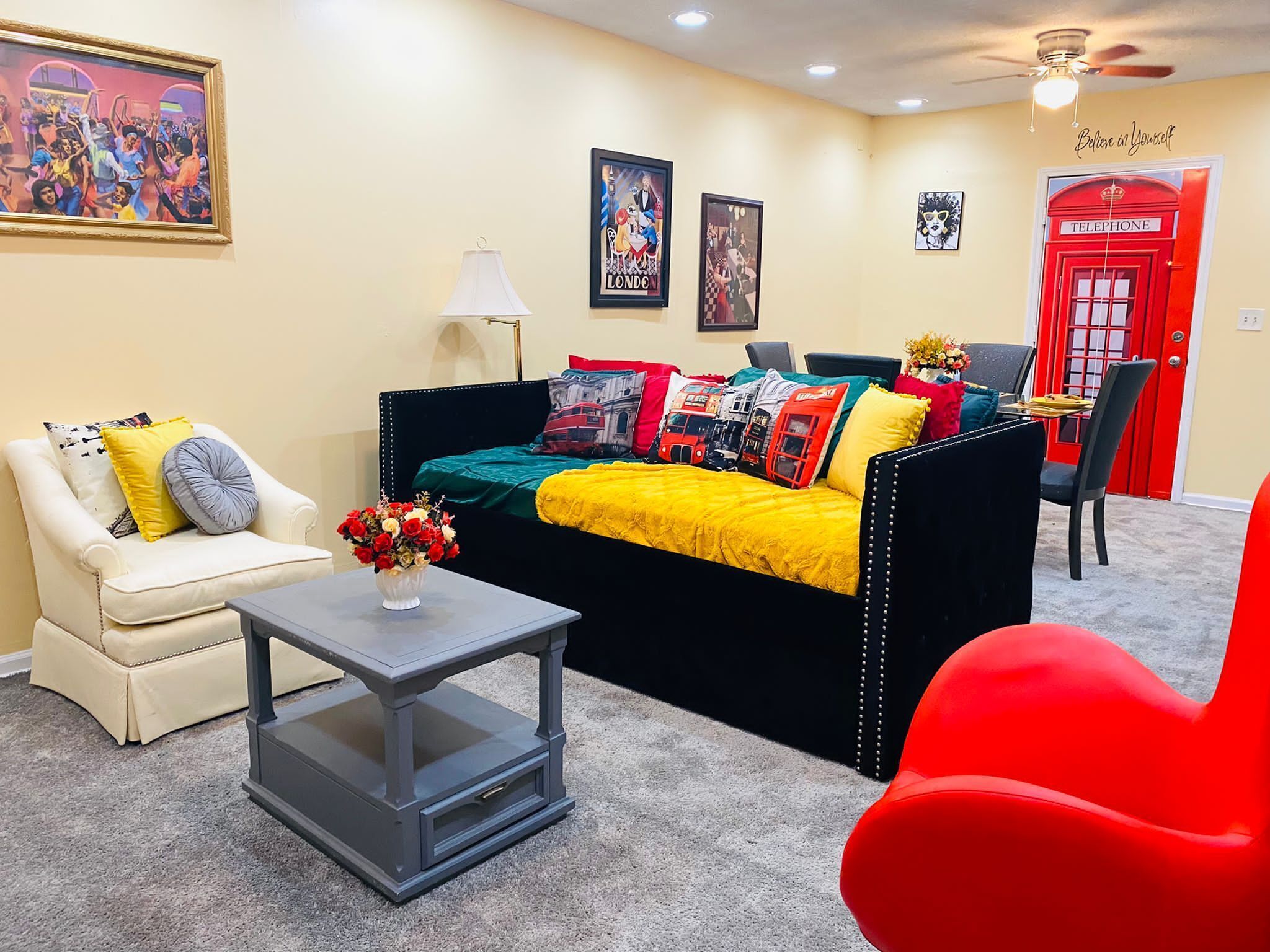 A living room with a black sofa, a white armchair, a red chair, a small table with a vase of flowers, and a lamp. There are posters on the wall and a red telephone booth decoration.