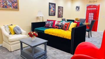A living room with a black sofa, a white armchair, a red chair, a small table with a vase of flowers, and a lamp. There are posters on the wall and a red telephone booth decoration.