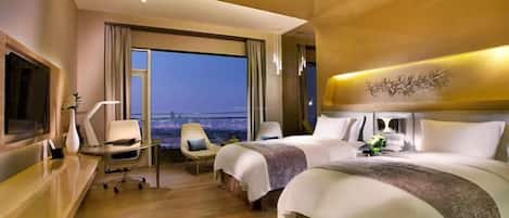 A hotel room with two beds, a desk, a chair, and a large window offering a city view.