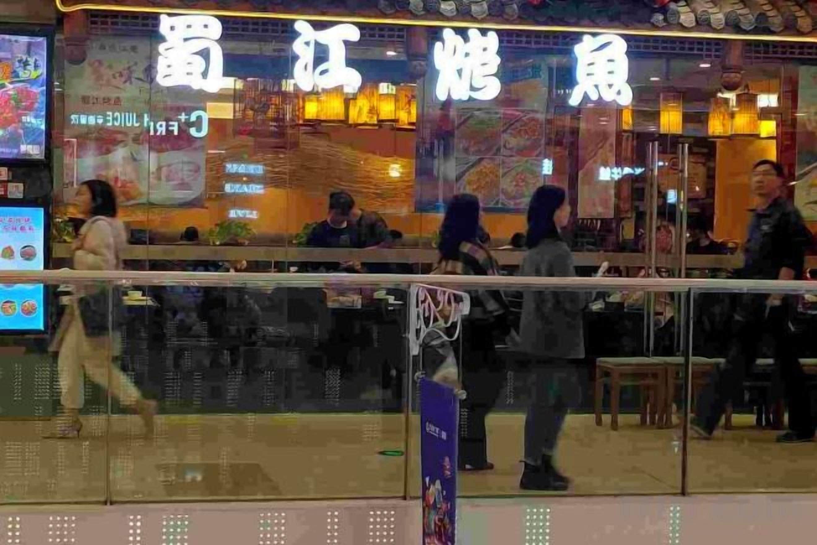 A restaurant with a glass barrier in front, people inside, and a digital menu display outside.