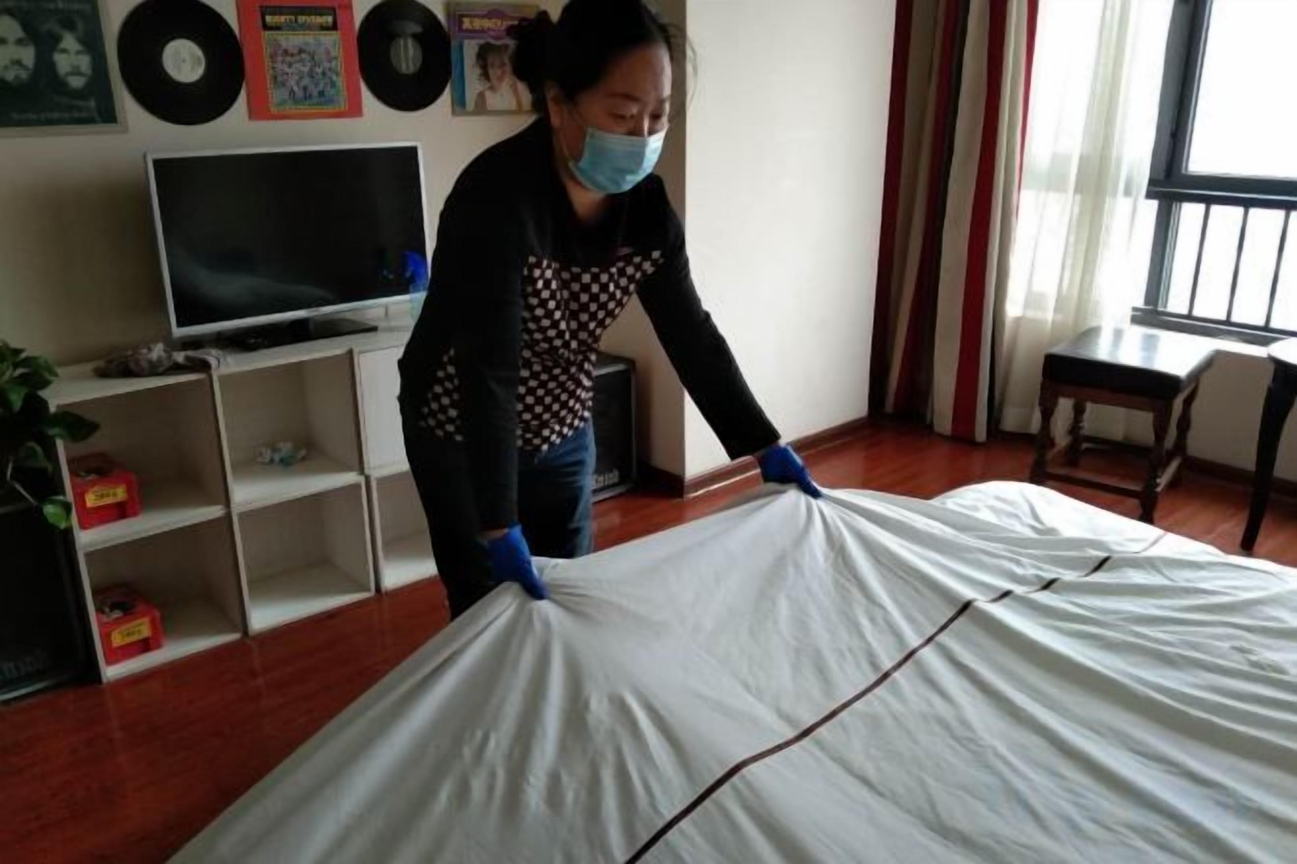 Una persona en una habitación con una cama cubierta de sábanas blancas, un televisor sobre un soporte y estantes con diversos objetos.