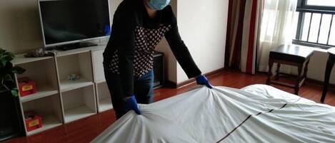 Una persona en una habitación con una cama cubierta de sábanas blancas, un televisor sobre un soporte y estantes con diversos objetos.