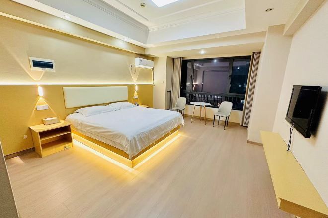 A modern hotel room with a large bed, bedside tables, a TV, a balcony with seating, and a view of the night sky.
