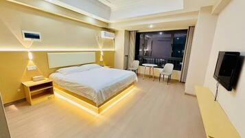 A modern hotel room with a large bed, bedside tables, a TV, a balcony with seating, and a view of the night sky.