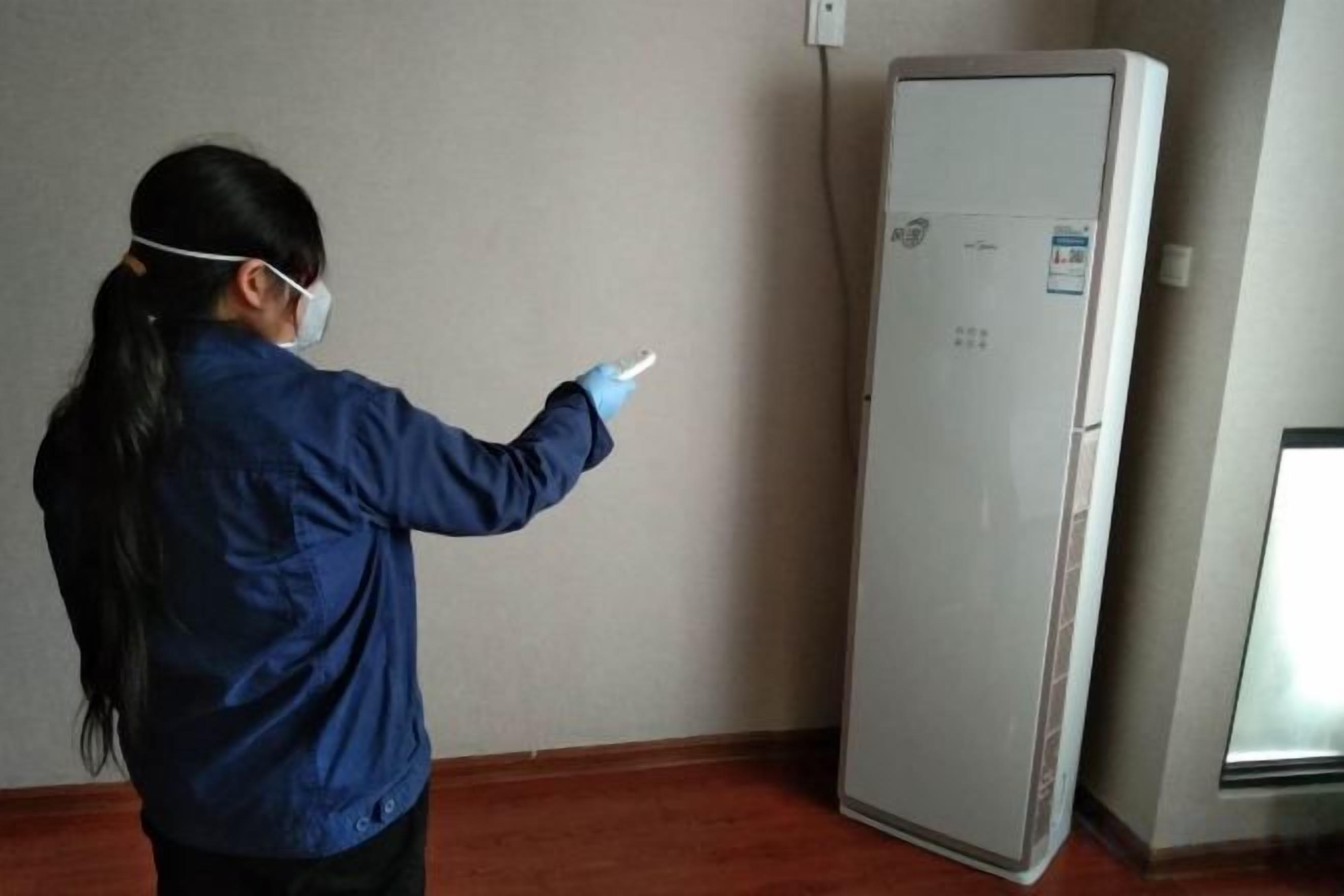 A person in a blue jacket interacting with a white appliance, possibly a refrigerator, in a room with a wooden floor and a plain wall.