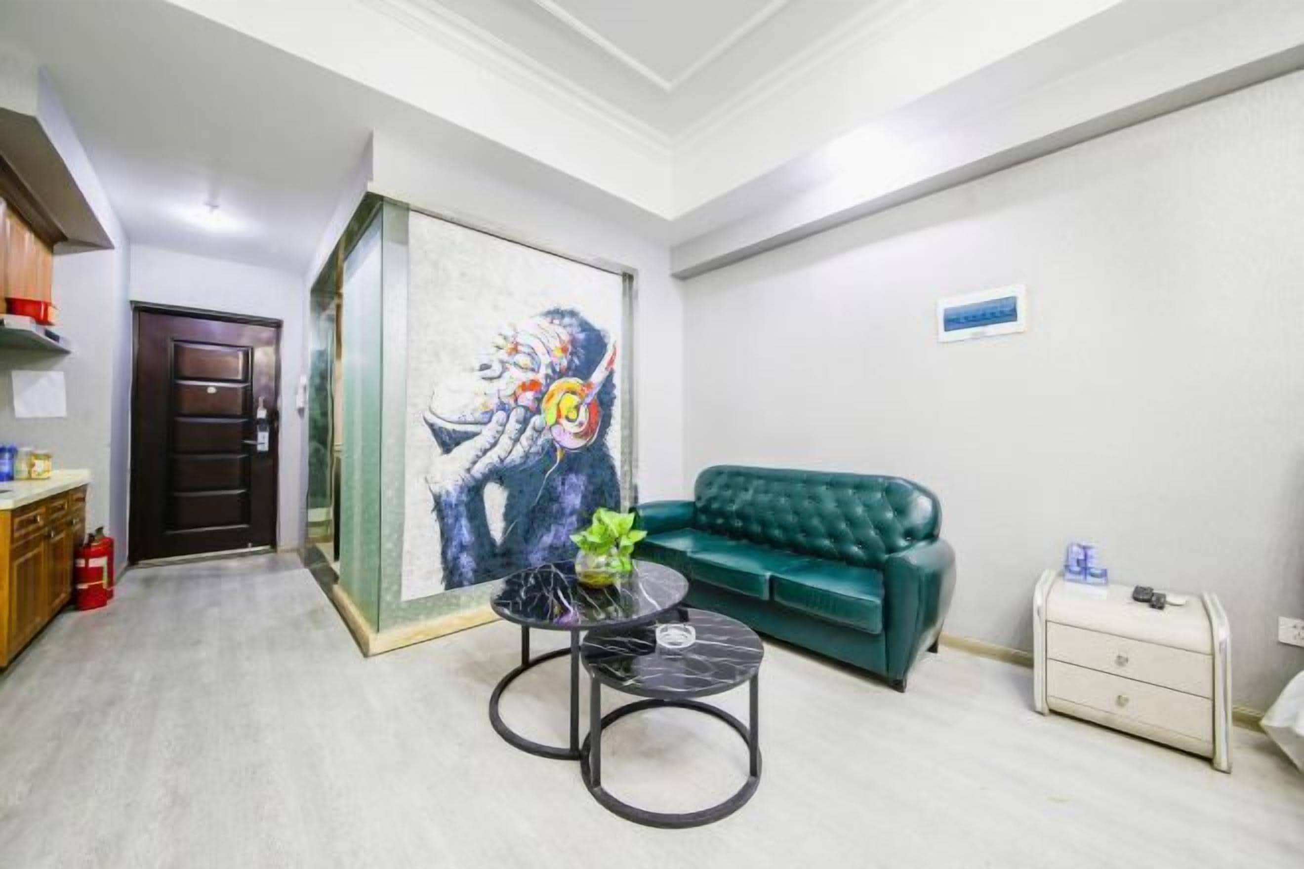 Una habitación con un sofá verde, una pequeña mesa redonda con un florero, un cuadro en la pared y una puerta con un extintor.