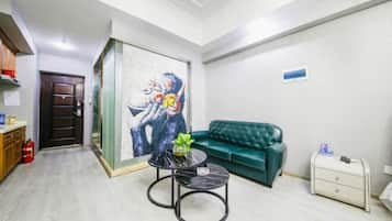 Una habitación con un sofá verde, una pequeña mesa redonda con un florero, un cuadro en la pared y una puerta con un extintor.