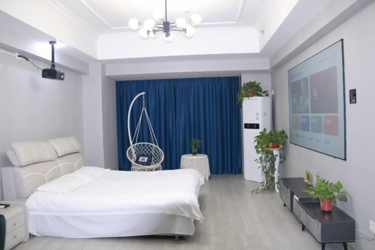 A modern hotel room with a large bed, a desk, a hanging chair, and a projector screen on the wall.