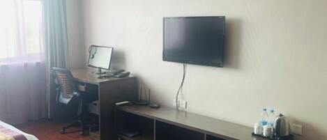 A hotel room with a bed, a desk with a computer, a television, and a shelf with water bottles.