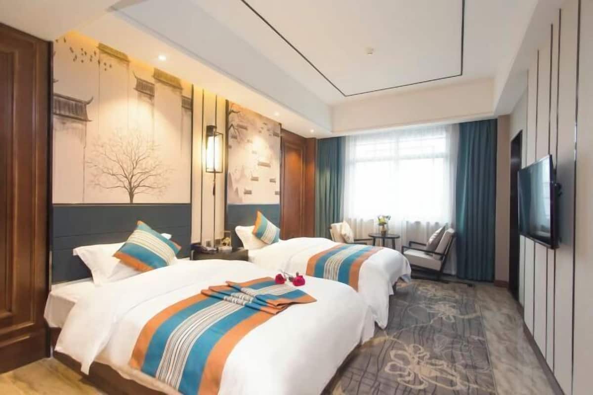 A hotel room with two beds, a TV, a desk, and a window with curtains.
