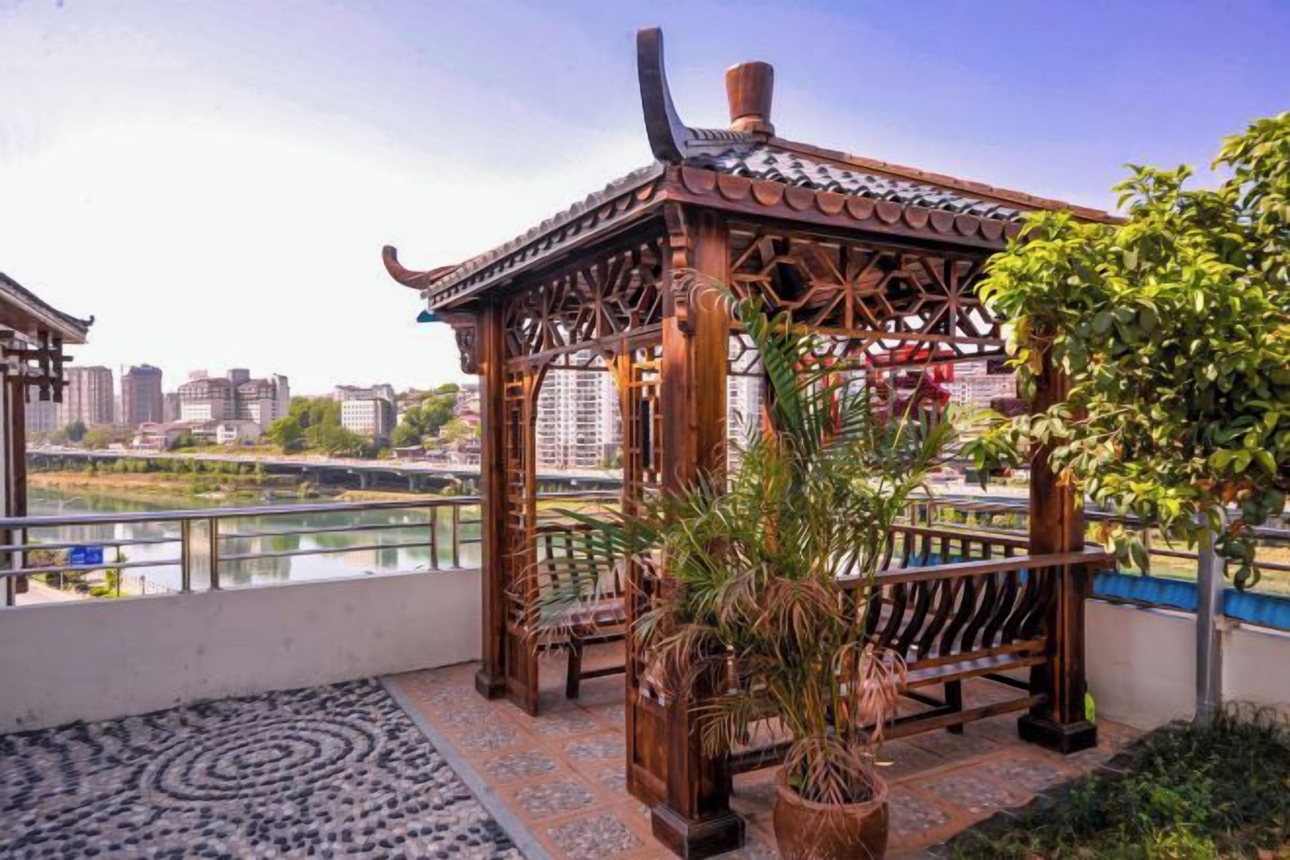 Un gazebo de madera tradicional con suelo de baldosas, una planta en maceta y vista a un río y edificios al fondo.