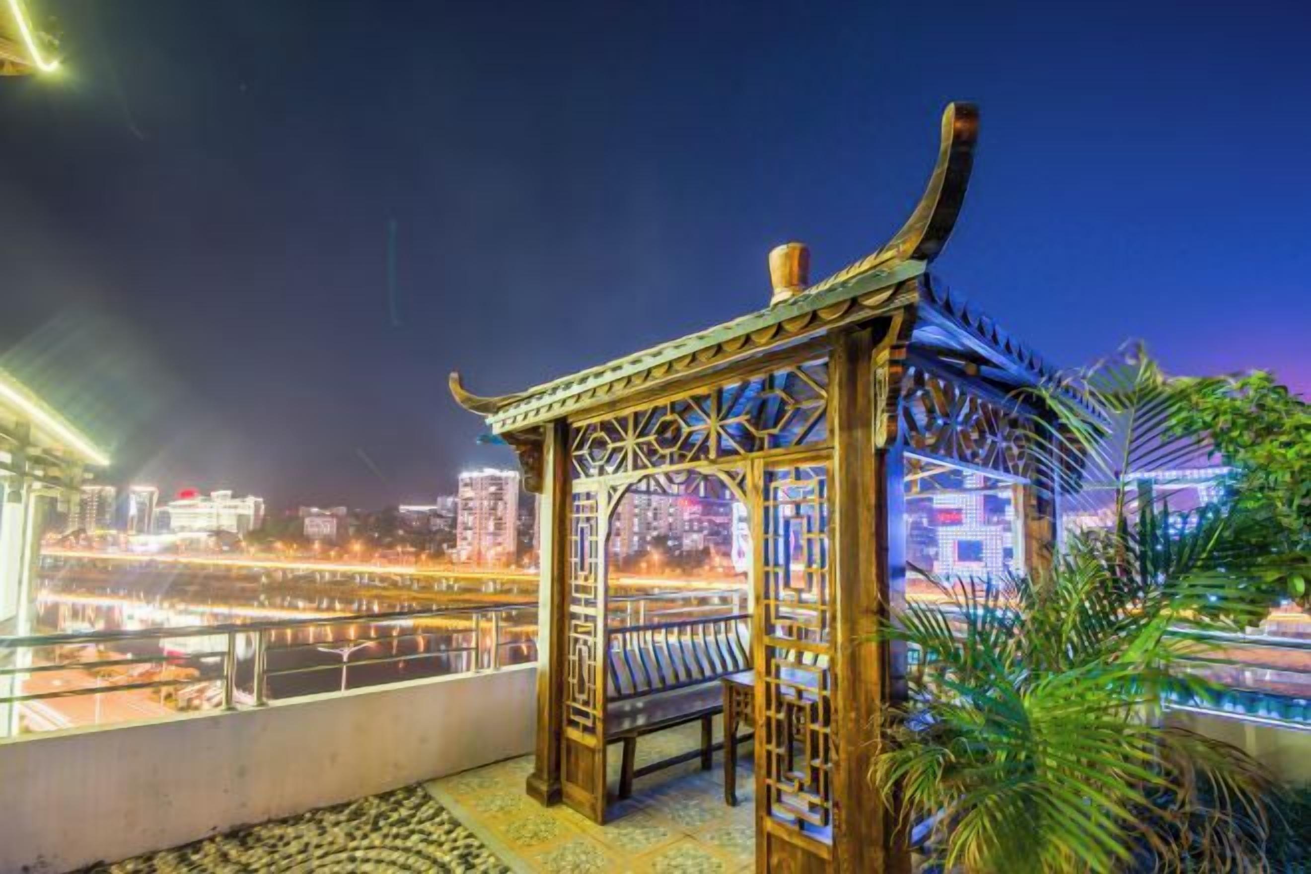 Un pabellón de madera tradicional con elaboradas tallas, con vistas a un paisaje urbano de edificios iluminados y un río.