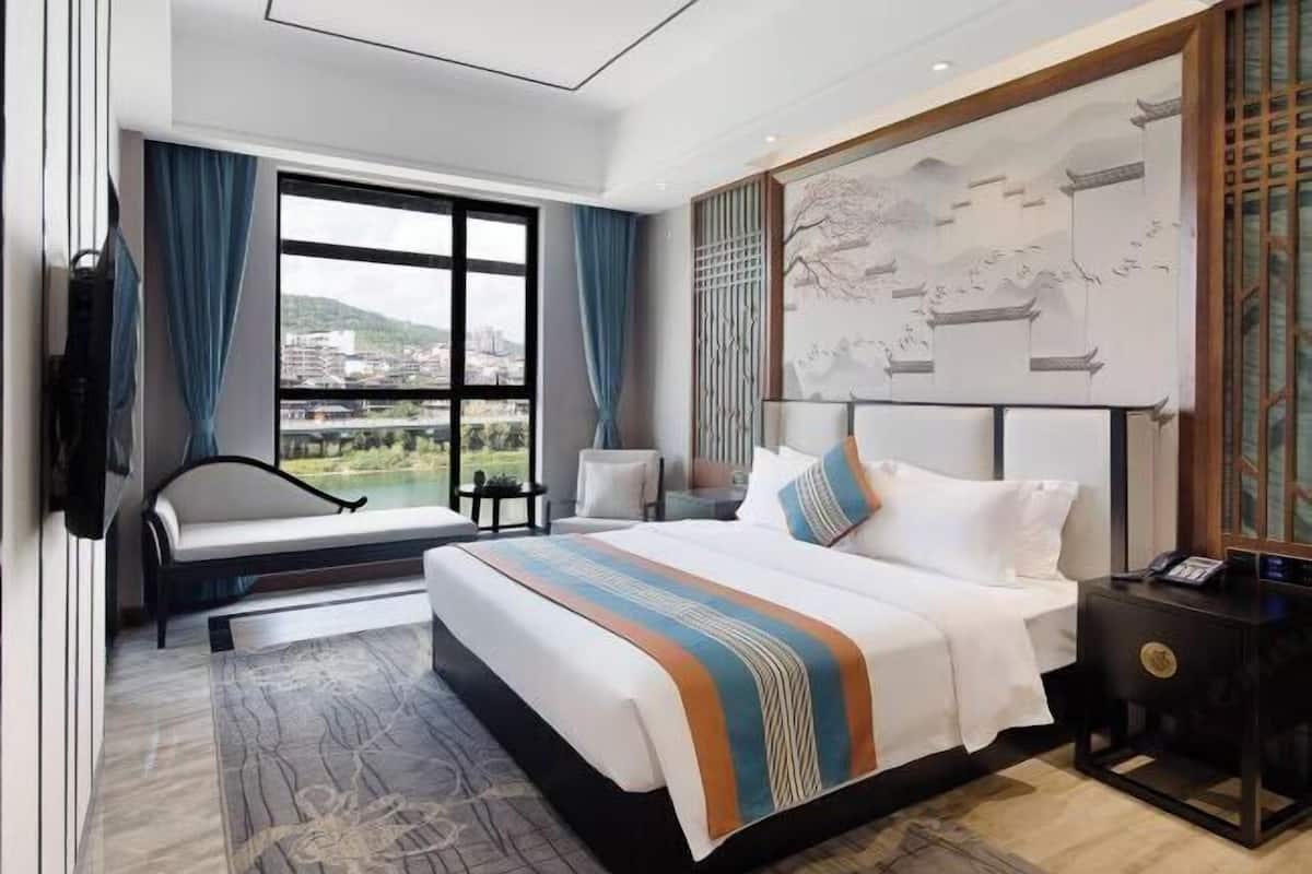 A modern hotel room with a large bed, a view of the city, a desk, and a chair.