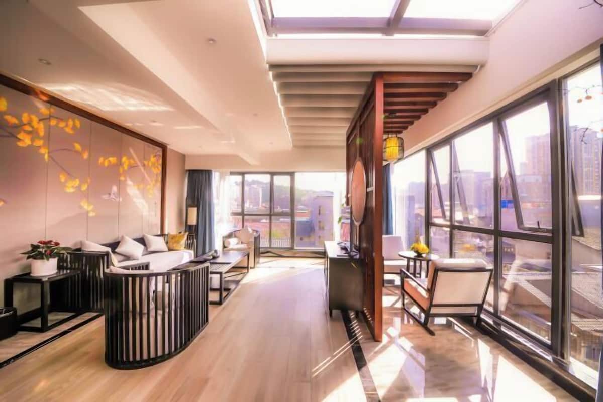 A modern hotel room with a large window, a sofa, a dining area, and a decorative wall.