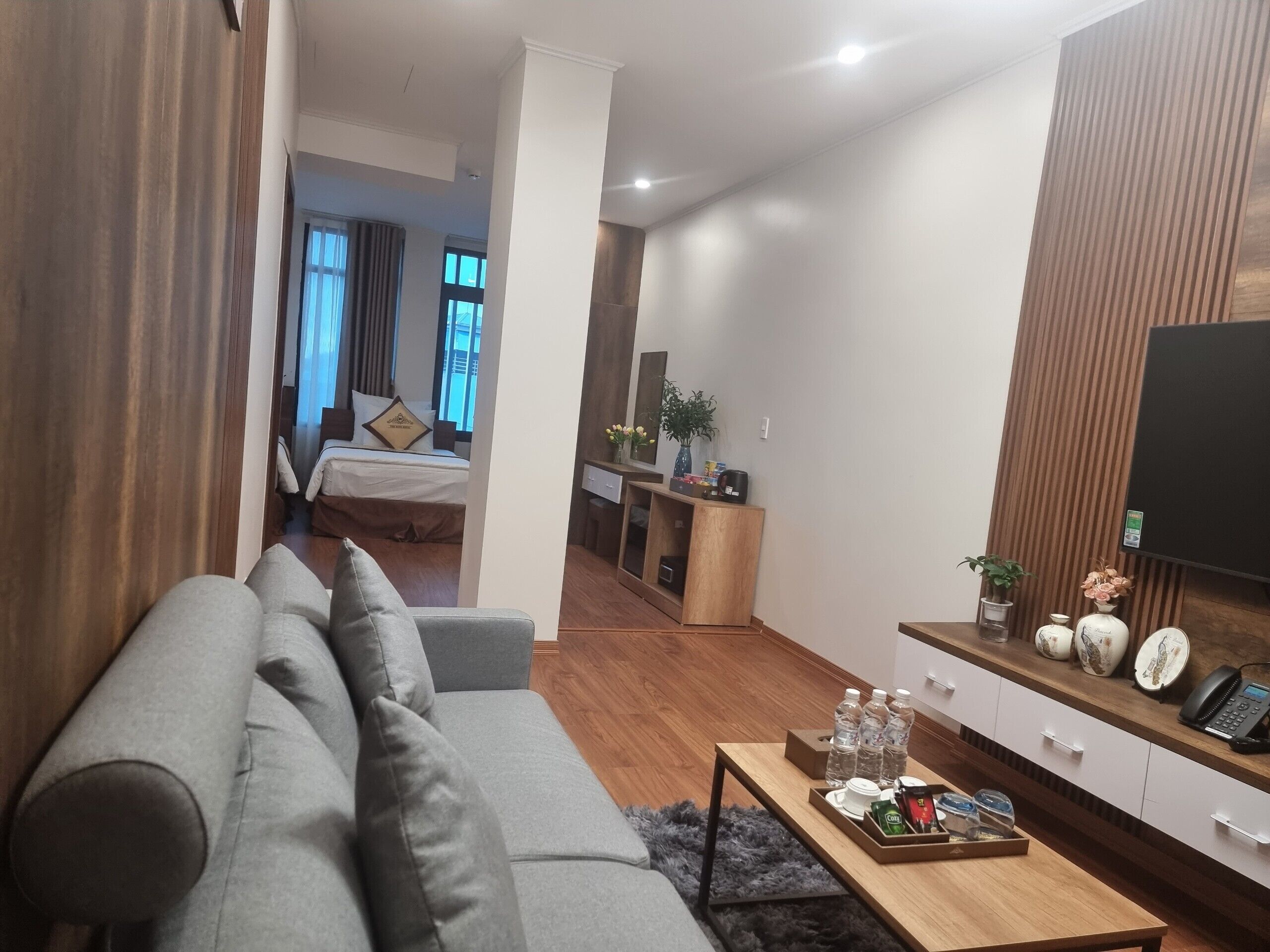 Ein modernes Wohnzimmer mit grauem Sofa, Holzboden, einem Couchtisch mit Annehmlichkeiten, einem Fernseher rechts und einem Schlafzimmer im Hintergrund.