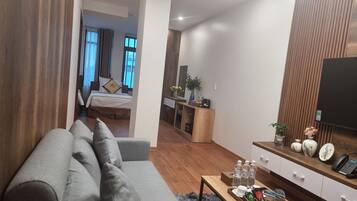 Ein modernes Wohnzimmer mit grauem Sofa, Holzboden, einem Couchtisch mit Annehmlichkeiten, einem Fernseher rechts und einem Schlafzimmer im Hintergrund.