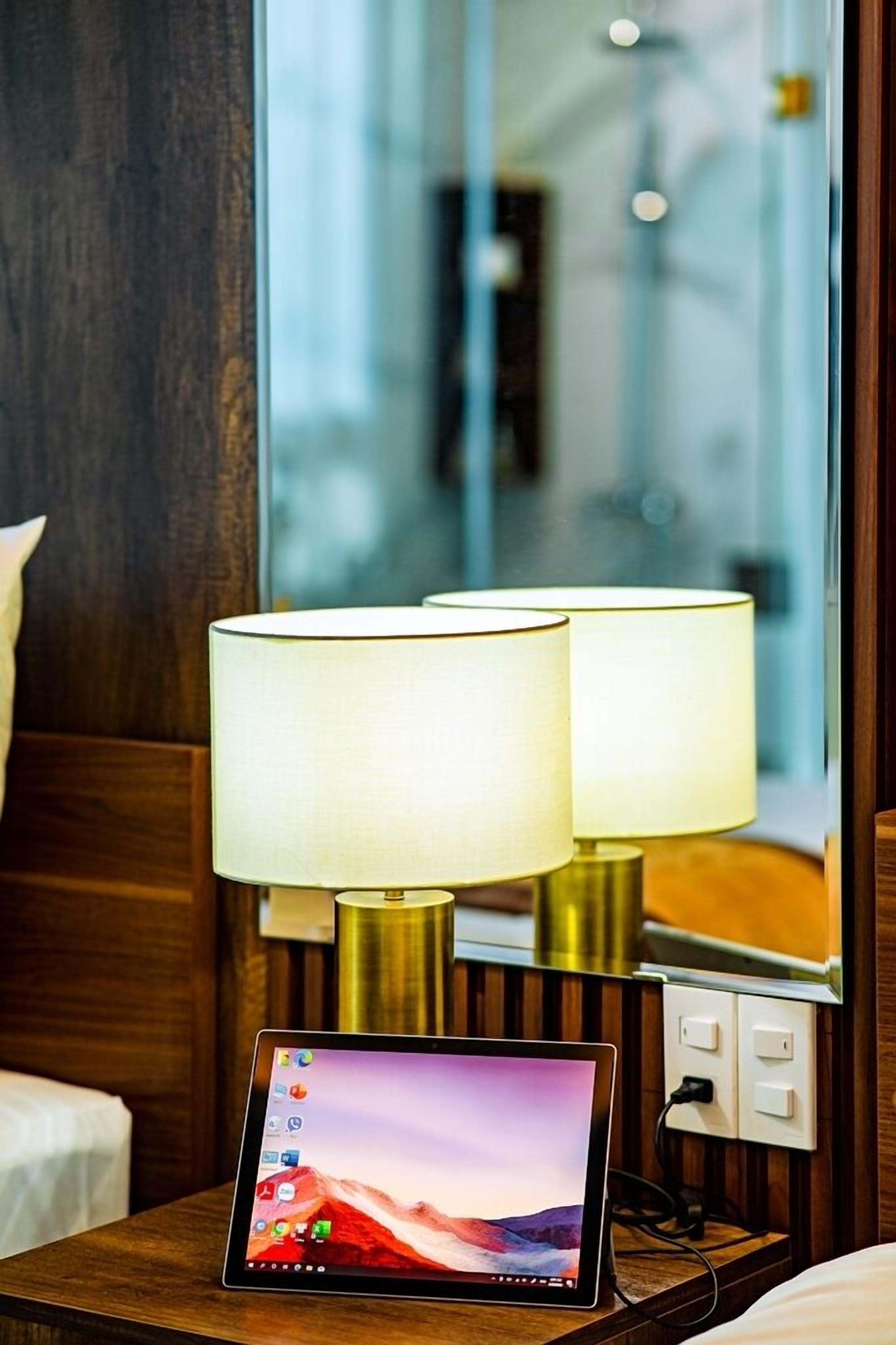A hotel room with a bedside table featuring a tablet, two lamps, and a mirror reflecting the room.